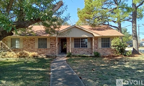 A house with a brown roof and a brick wall is surrounded by green grass and trees.