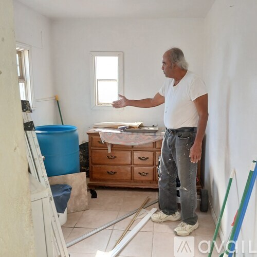 A man in a white shirt and jeans is standing in a room with a blue bucket and a wooden dresser.
