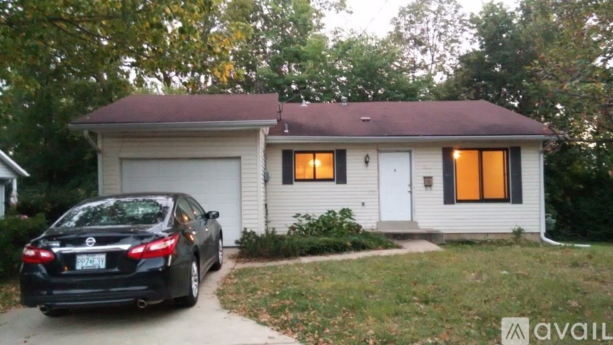 A black car is parked in front of a white house with orange windows.