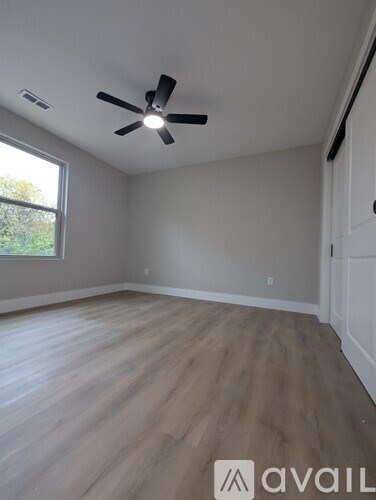 A room with a ceiling fan and light fixture, wooden flooring, and a window with a view of trees outside.