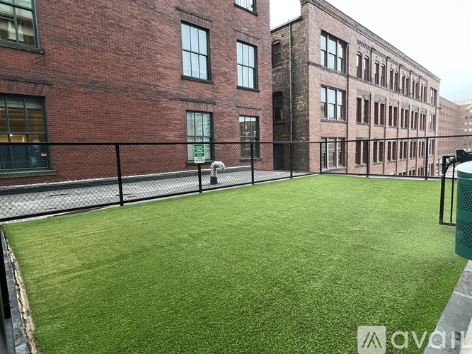 A green artificial turf area is surrounded by a black fence and two brick buildings.