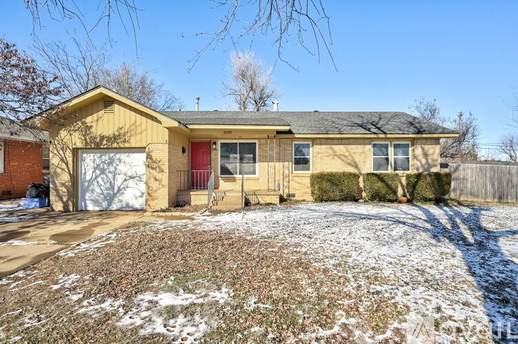 A house with a red door and a grey roof is surrounded by a snowy ground.