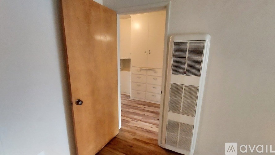 A room with a wooden door leading to another room with a white wall and a white air conditioner unit.
