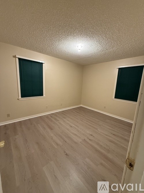 A room with light wood flooring and two windows with black blinds.