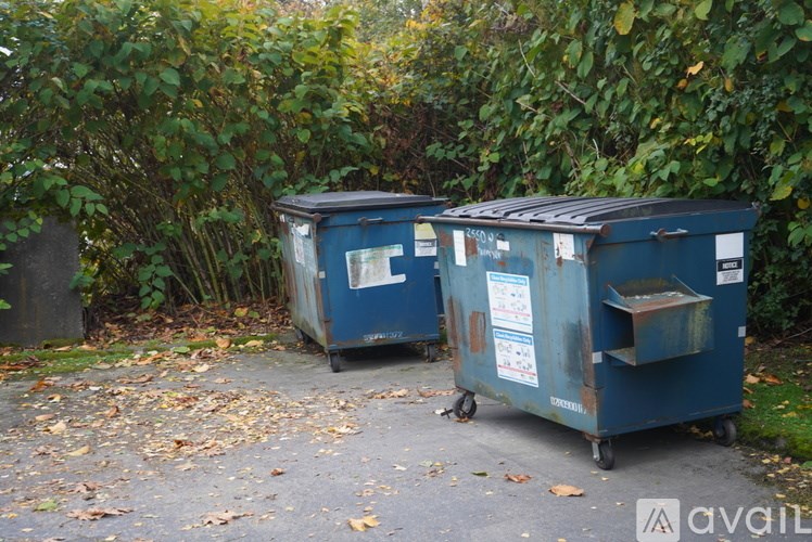Two blue dumpsters are parked next to a tree.