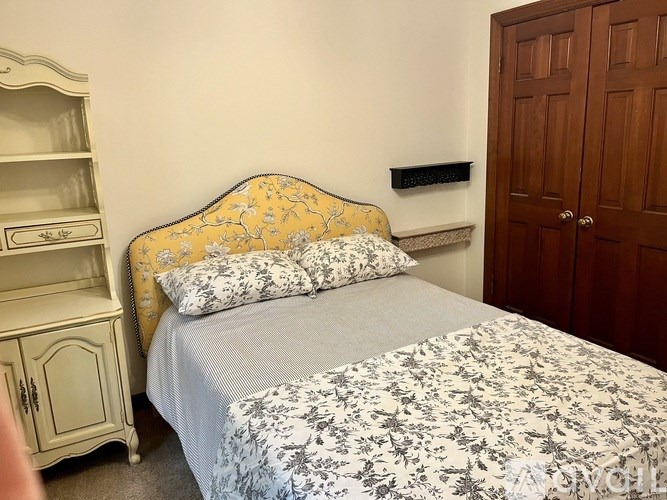 A bed with a floral patterned cover and a yellow headboard sits in a room with a wooden door and a white nightstand.