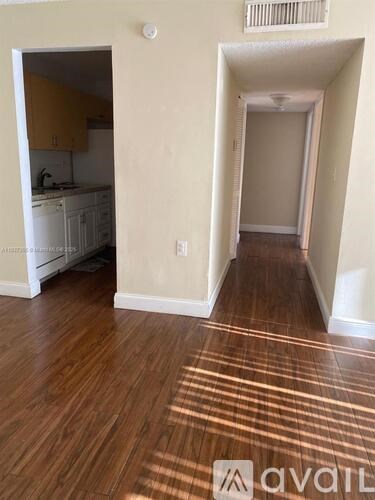 A room with wooden floors and a kitchen area in the background.