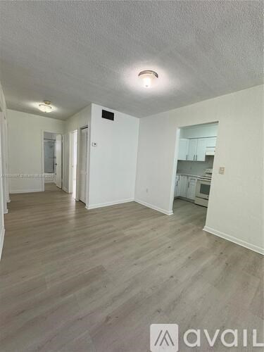 A room with wooden flooring and white walls, with a kitchen visible in the background.