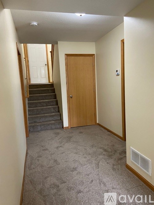 A hallway with a carpeted floor, a staircase with a white door, and a wooden door at the end of the hallway.