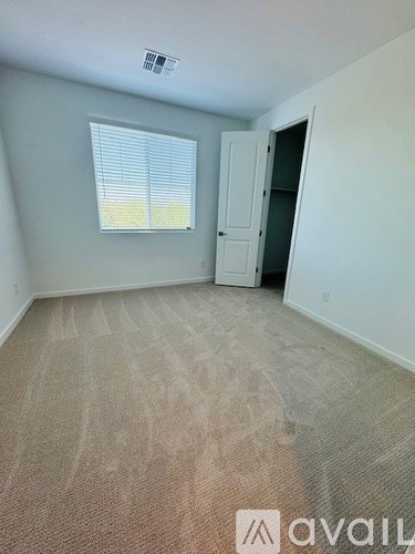 A room with a carpeted floor, a window with blinds, and a door leading to another room.