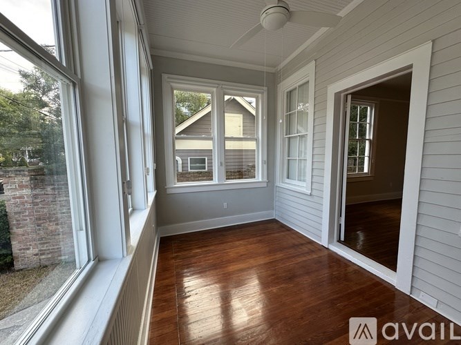 A room with wooden floors and white walls, with a ceiling fan and a view of a house through the windows.
