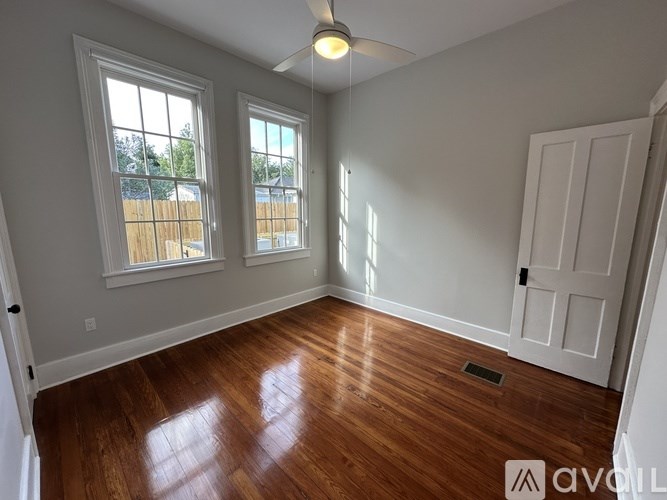 A room with wooden floors and white walls has a ceiling fan and two windows.