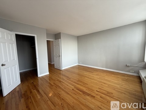 A room with wooden flooring and white walls, with a door on the left and a window on the right.