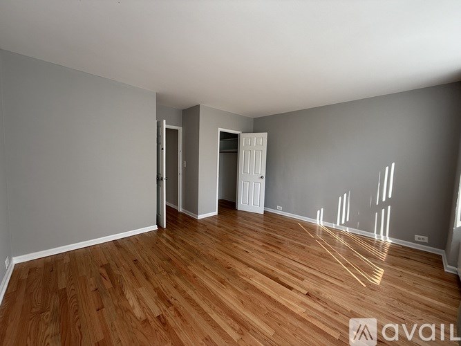 A room with wooden flooring and white walls, with a door on the left and a window on the right.