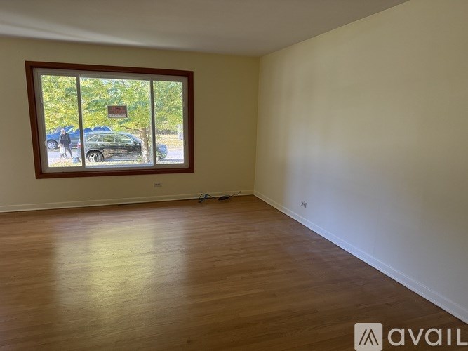 A room with wooden flooring and a window showing a parking lot outside.