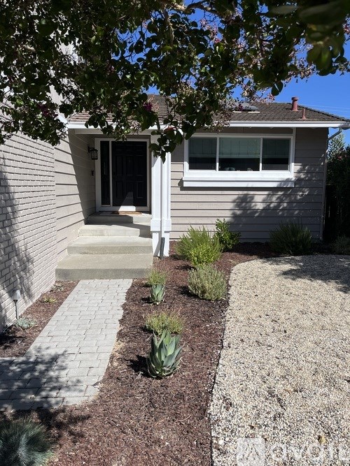 A house with a grey siding and a white door is surrounded by a garden with a gravel path.