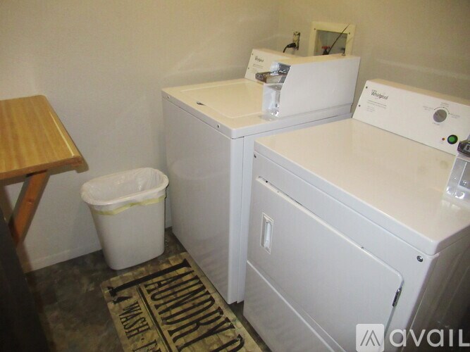 A white washer and dryer in a small room with a trash can and a wooden stool.