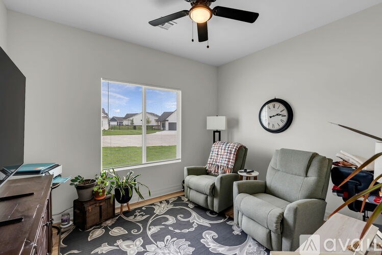 A living room with a ceiling fan, two chairs, a table, and a window with a view of houses outside.