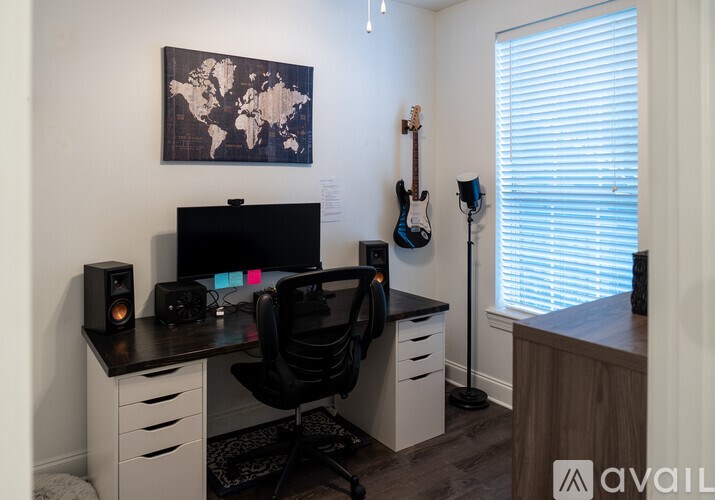 A black chair is in front of a desk with a computer and a guitar hanging on the wall.