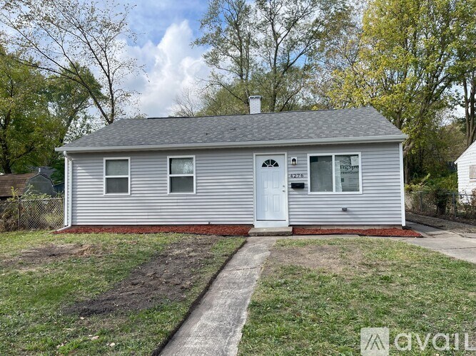 A small house with a grey siding and a white door is surrounded by a grassy area and trees.