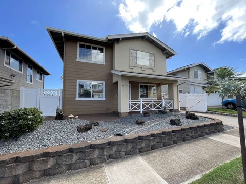 A house with a brown siding and a white door is surrounded by a gravel area and a small garden.