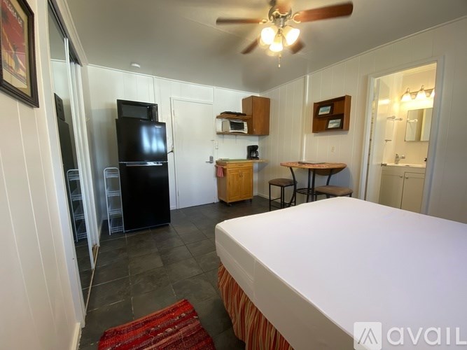 A bedroom with a bed, a table, chairs, a refrigerator, and a ceiling fan.