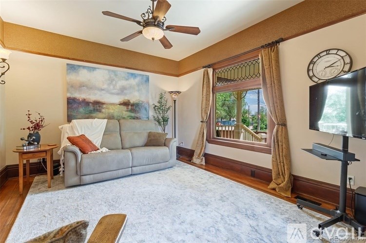 A living room with a grey couch, a ceiling fan, a painting on the wall, a window with curtains, and a television.