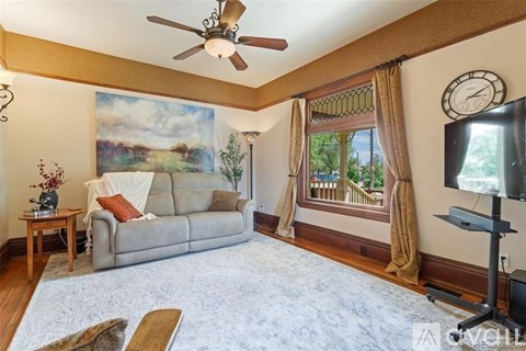 A living room with a grey couch, a ceiling fan, a painting on the wall, a window with curtains, and a television.