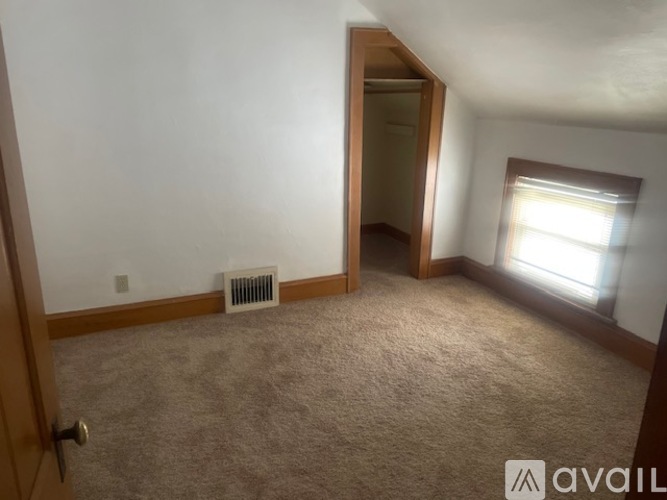 A room with a carpeted floor, a window with blinds, and a door leading to another room.