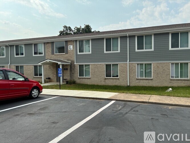 A red car is parked in a parking lot in front of a grey apartment building.
