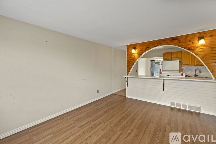 A room with wooden flooring and a white wall with a wooden archway leading to a kitchen area.