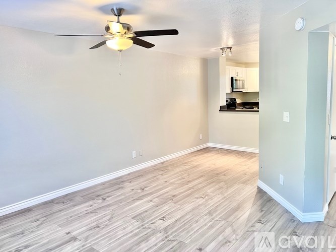 A room with a ceiling fan and light fixture, hardwood flooring, and a small kitchenette in the background.
