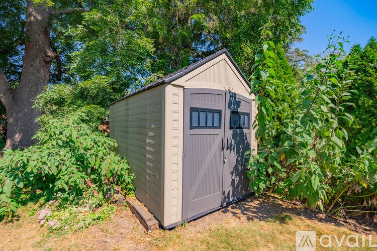 A grey shed with a brown roof is surrounded by green plants.