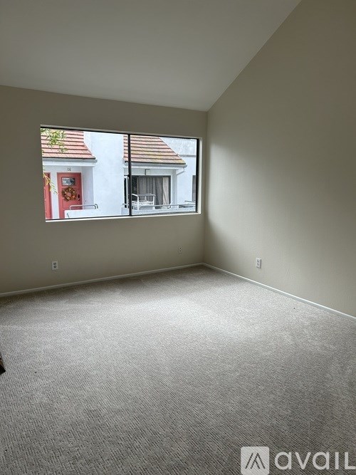 A room with a carpeted floor and a window showing a red door and a balcony.