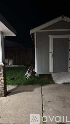 A small white shed with a door is situated in a yard.