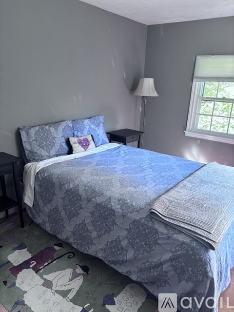 A bed with a grey and blue comforter and a white pillow with a purple heart on it.