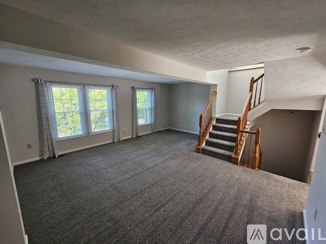 A spacious room with a carpeted floor, a staircase with wooden railings, and a window with curtains.
