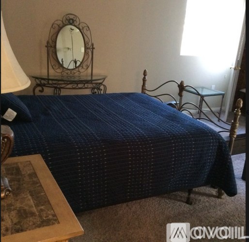 A bed with a blue comforter sits in a room with a mirror and a nightstand.