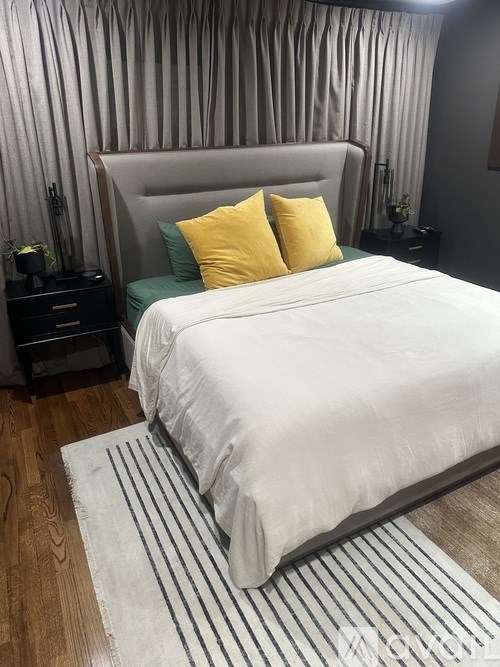 A bed with a white comforter and two yellow pillows sits in a room with a striped rug in front of it.