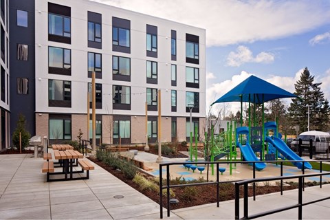 A playground with a blue umbrella and a slide is in front of a building.