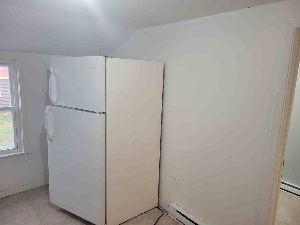 A white refrigerator stands in a room with a window to the left.