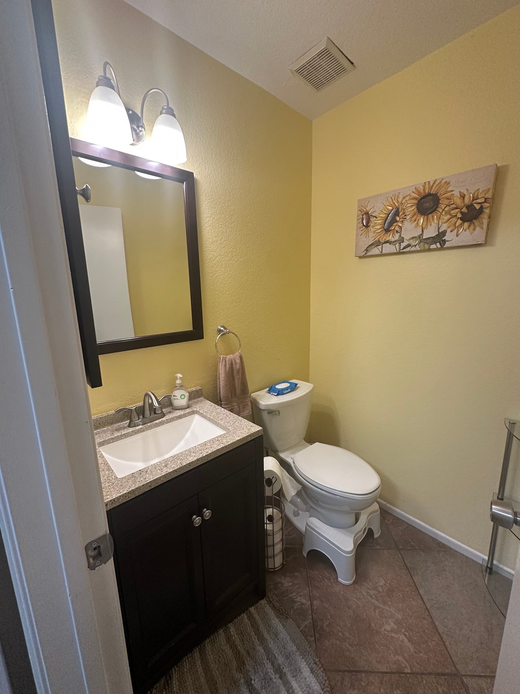 A bathroom with a toilet, sink, mirror, and a sunflower picture on the wall.