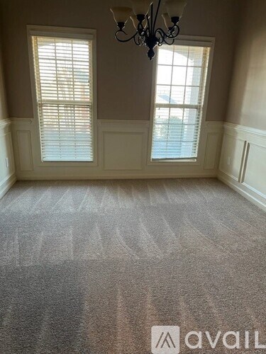 A room with a carpet, a chandelier, and two windows with blinds.