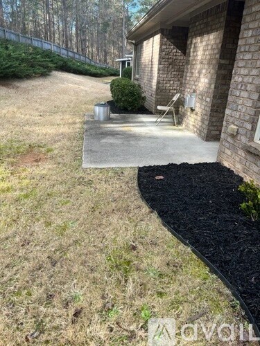 A concrete patio leads to a brick house with a black mulch garden bed.