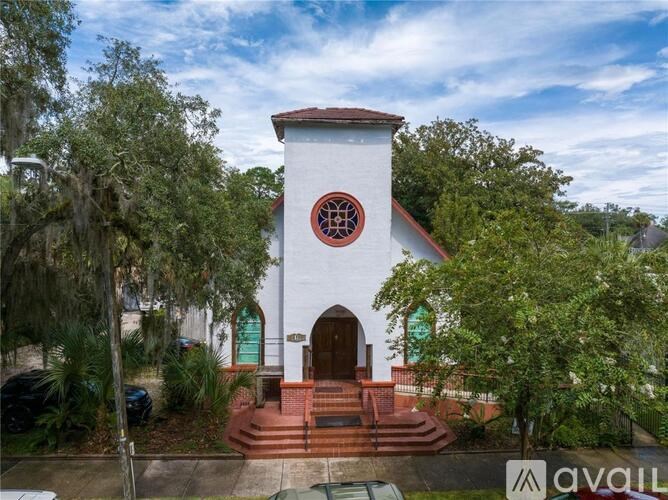 A white church with a red roof and a circular window is surrounded by trees.