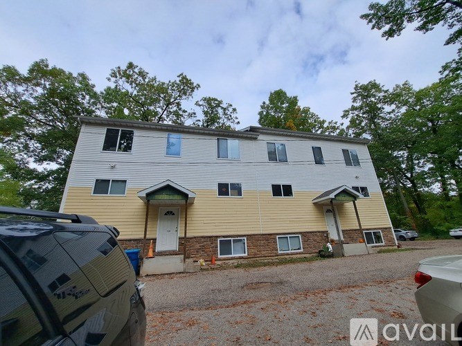 A two-story house with a beige and brown exterior is surrounded by trees and has a car parked in front.