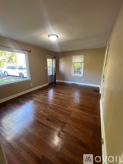 A room with wooden flooring and a window with a view of a car and trees outside.