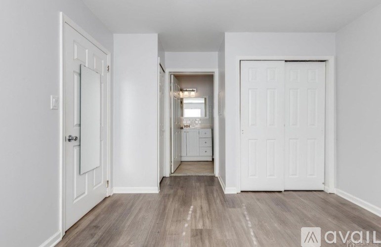 A room with white walls and wooden flooring, leading to a hallway with a light on.