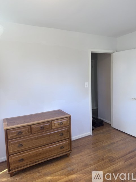 A wooden chest of drawers sits in a room with a white wall and a doorway leading to another room.