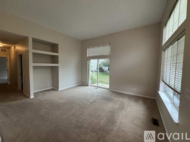 A spacious room with a carpeted floor, a window with blinds, and a door leading to another room.